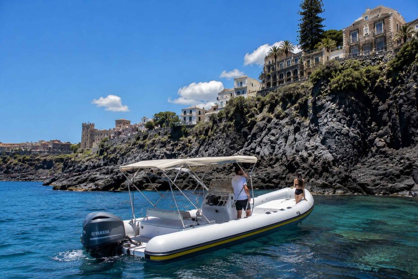 A white inflatable boat with a Yamaha motor on calm blue water, a rocky coastline, and historic buildings in the background. 2