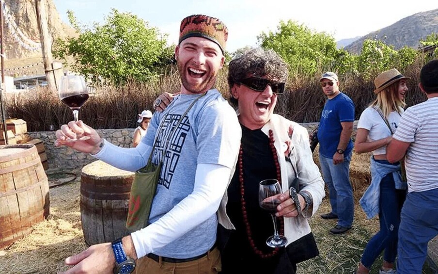 People standing outdoors, holding wine glasses, with wooden barrels and trees in the background. 1