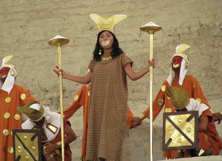 A person in a patterned dress holding a staff, flanked by two individuals in orange robes with gold accents. 3