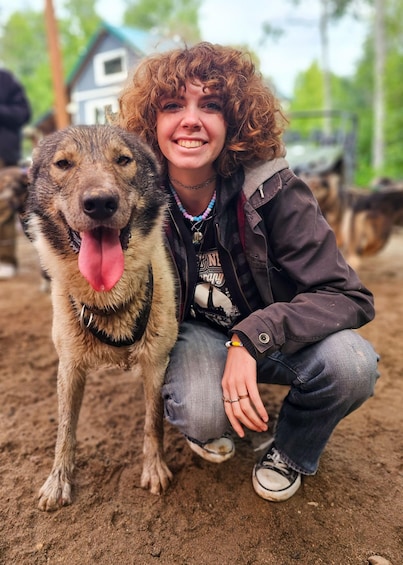 A person crouching next to a dog on sandy ground with a house and trees in the background. 1