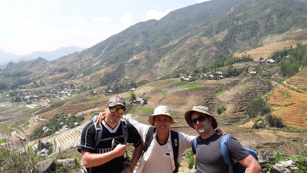 Three people standing on a mountain ridge overlooking terraced fields and a village. 5