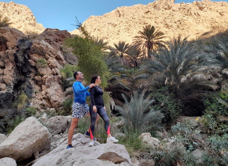 Two people standing on a rocky outcrop with palm trees and a rocky mountain in the background. 2