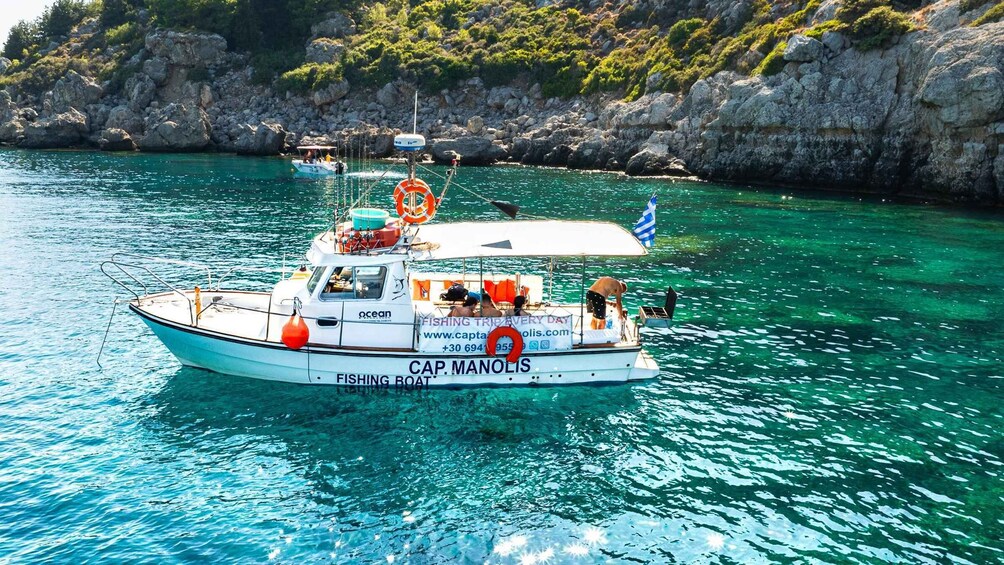 A fishing boat named CAP. MANOLIS in clear turquoise waters with rocky cliffs in the background. 1