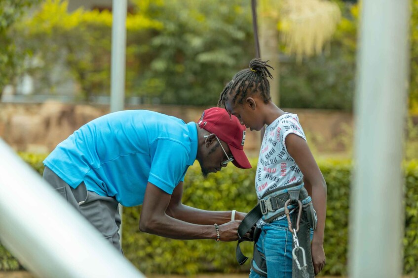 A person in a blue shirt and red cap assisting another person in a white shirt with red text and blue jeans, both wearing safety harnesses. 1