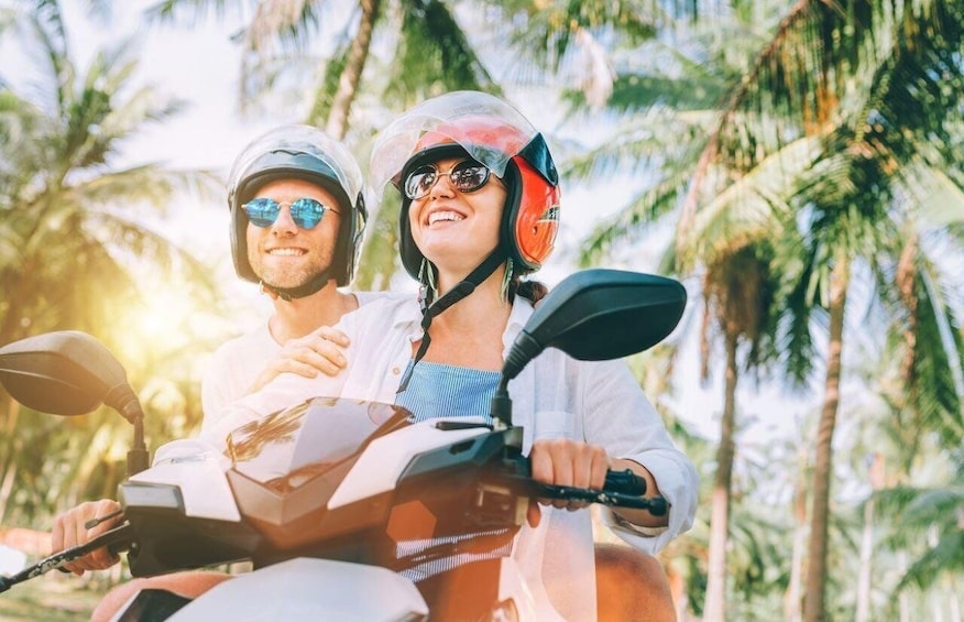 Two people on a motorcycle with helmets, surrounded by palm trees. 1