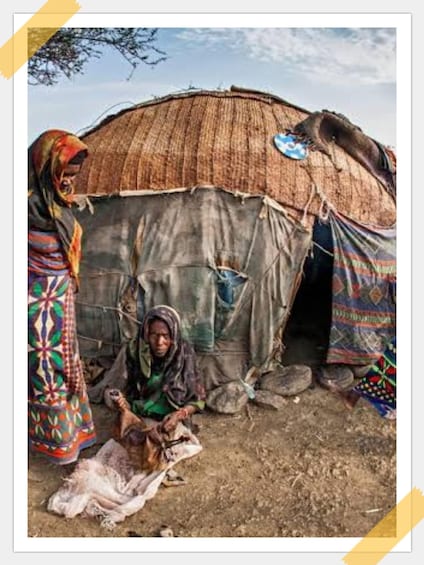 A thatched hut with two women, one sitting and one standing, in front of it. 1
