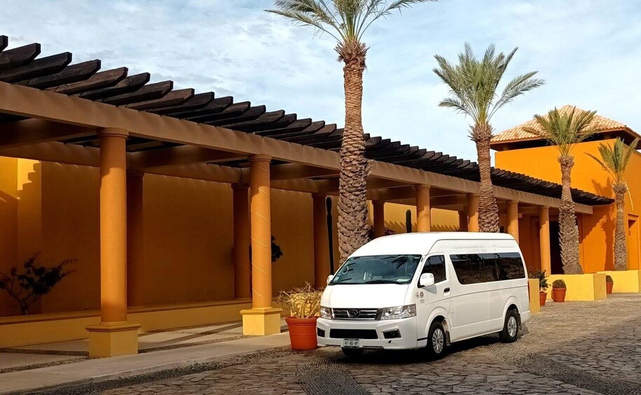 A white van parked in a courtyard with a pergola, palm trees, and a yellow building. 2