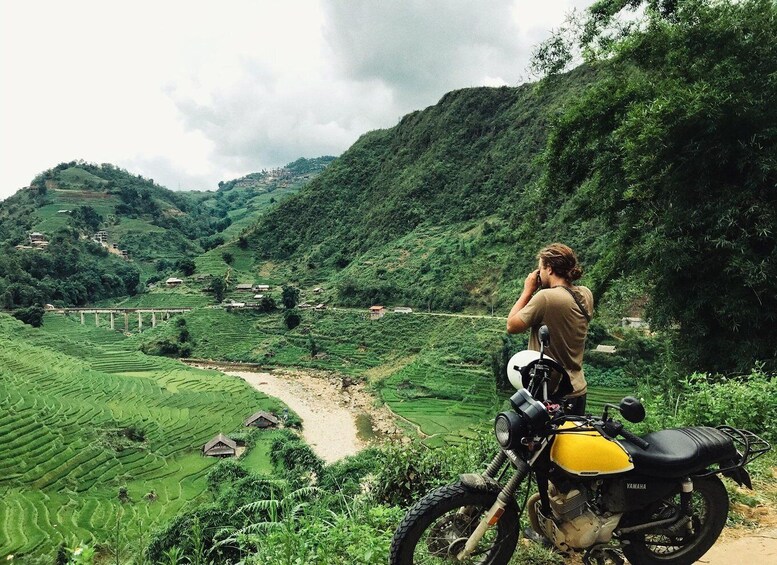 A person standing next to a yellow motorcycle overlooking terraced fields and a bridge in a hilly landscape. 5