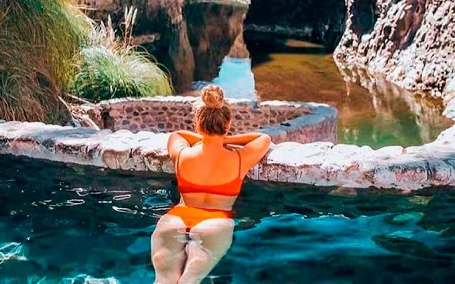 A person in a swimsuit sitting by a clear pool with a rocky edge and greenery in the background. 1