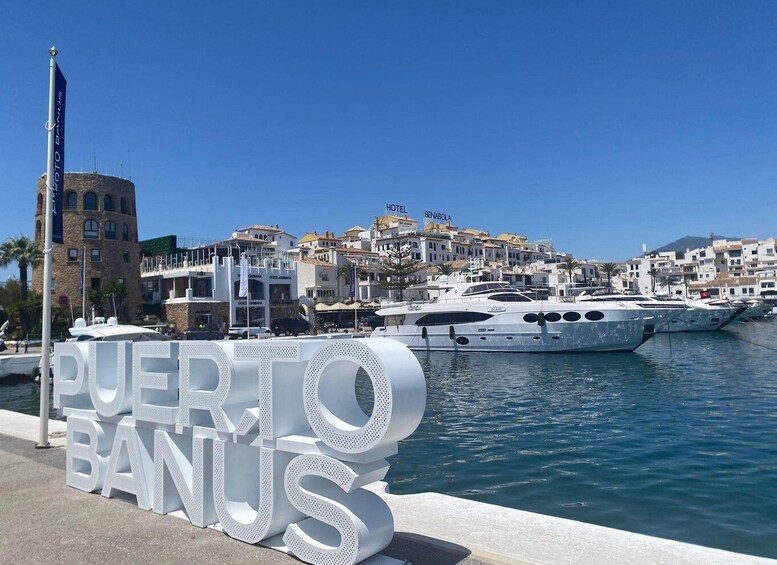 A marina with yachts, a large sign reading "PUERTO BANUS", and buildings in the background. 1