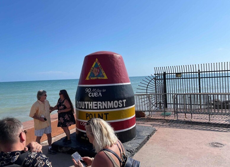 A large, colorful marker with '90 Miles to Cuba' and 'Southernmost Point' written on it, located near a beach with a metal fence and people standing nearby. 2
