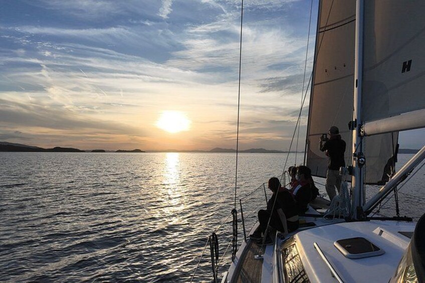A sailboat at sunset with people on deck, calm water, and a distant shoreline. 1