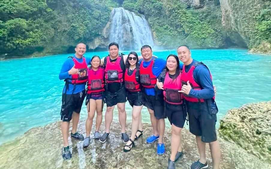 A group of people standing on a rock by a waterfall in a turquoise body of water. 5