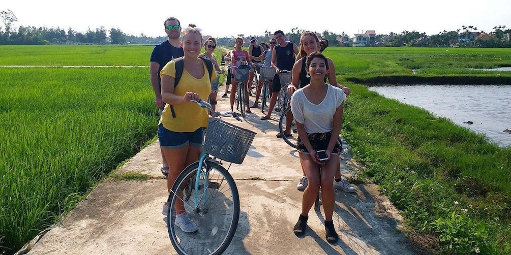 A group of people walking and cycling along a concrete path beside a waterway, with green fields and trees in the background. 2