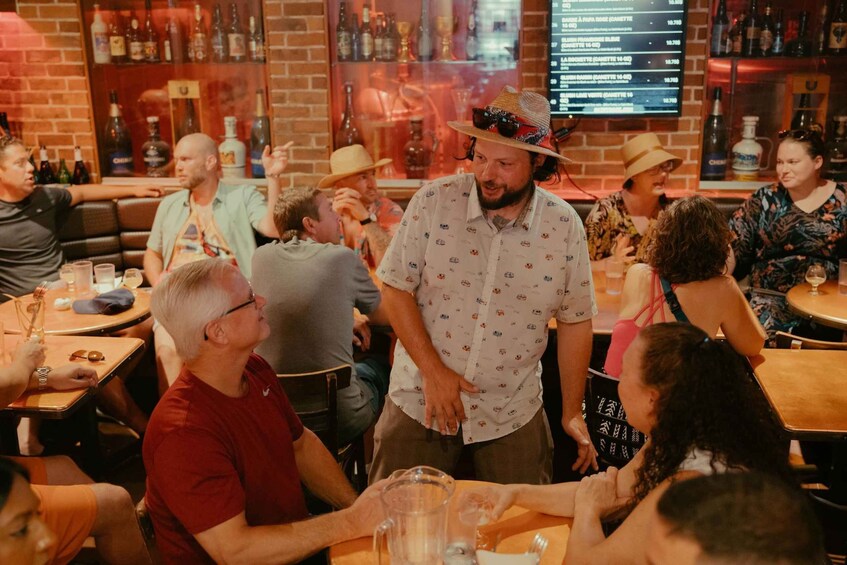 A group of people in a bar with tables and chairs, bottles on shelves, and a menu on the wall. 1