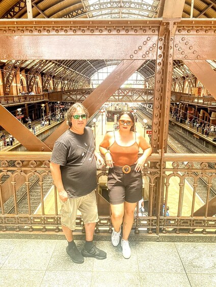 A man and a woman standing on a bridge with a metal railing. The bridge has a high ceiling with a glass roof and a metal framework. There are people walking on the bridge and a train track below. 4