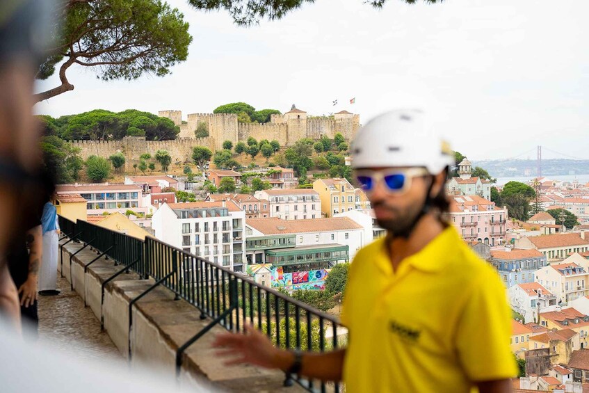 A person in a yellow shirt and helmet on a balcony overlooking a historic city with a prominent castle and colorful buildings. 2