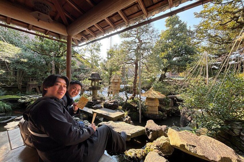 Two people sitting on a wooden bench in a garden with a pond, surrounded by rocks and plants. 4