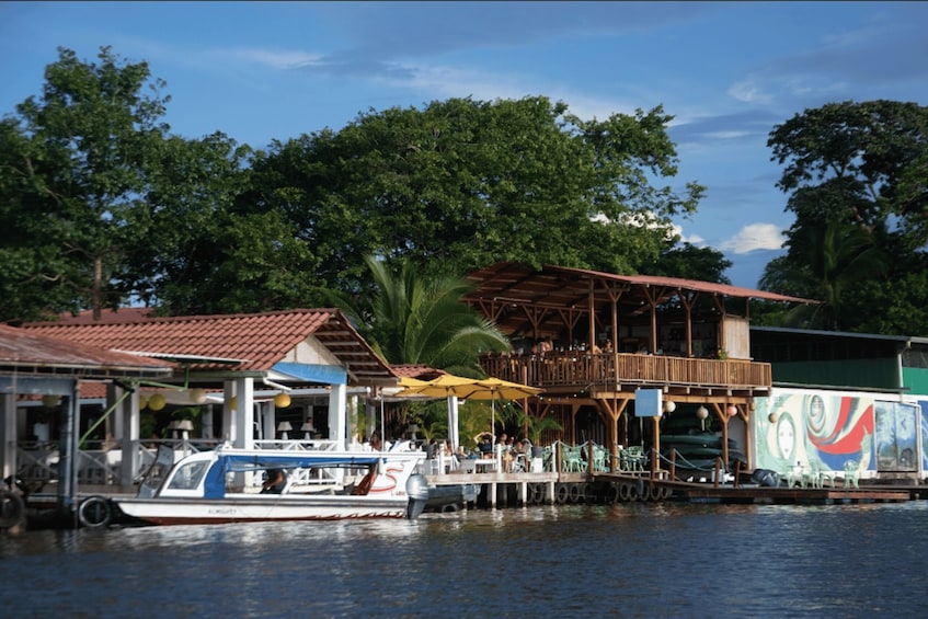 A boat docked at a waterfront restaurant with outdoor seating and a mural. 2