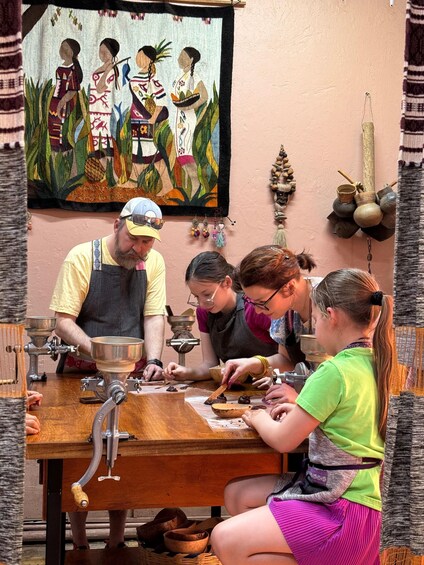 A group of people engaged in a craft activity, possibly pottery or cooking, with a painting of indigenous figures on the wall. 4