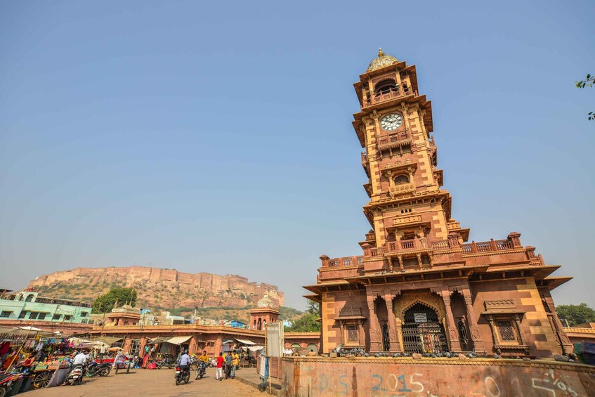 A historic clock tower with intricate architecture, a bustling market area with people and vehicles, and a large fort in the background. 3