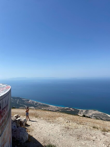 A person standing on a rocky hillside overlooking a coastal landscape with a clear sky. 2