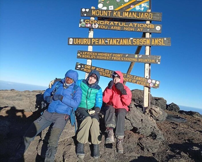 Three people sitting on a rocky outcrop with a signpost in the background. 4