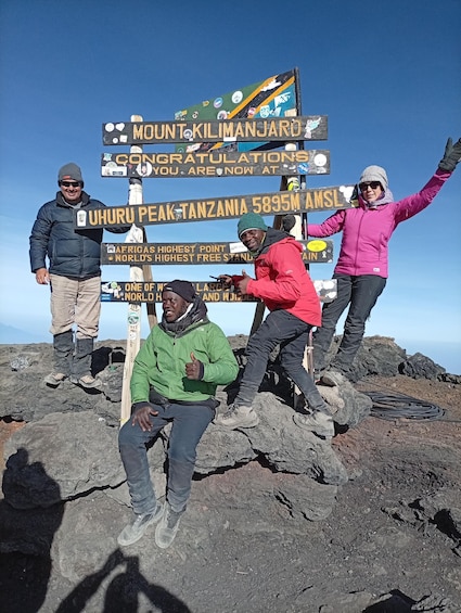 A group of people at Uhuru Peak, Tanzania's highest point, with a signboard displaying various facts about the location. 1