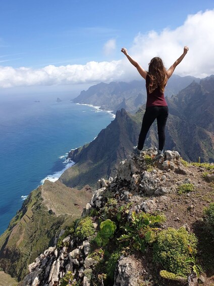 A woman standing on a rocky cliff with arms raised, overlooking a coastal landscape with mountains and the ocean. 3