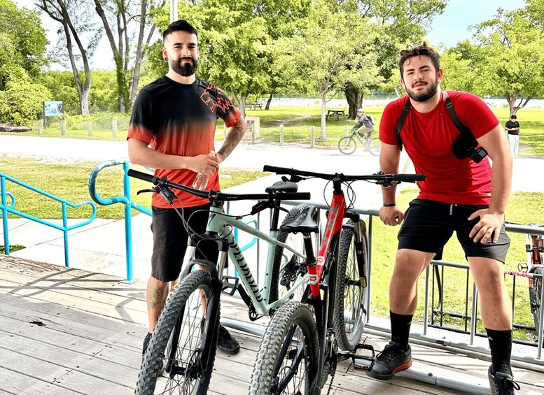 Two men standing next to bicycles on a wooden deck in a park. 2