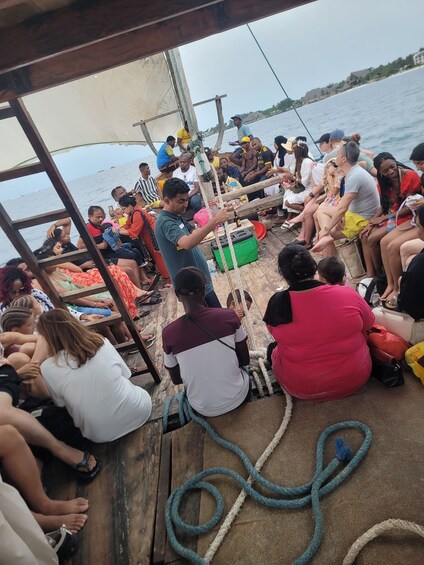 A group of people on a boat with a sail, some sitting and others standing, near a body of water. 2