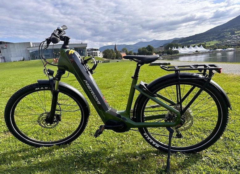 A green electric bike with a rear rack on a grassy field with buildings and mountains in the background. 1
