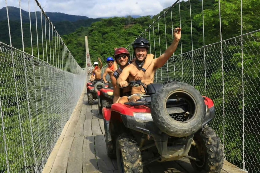 A group of people on ATVs on a suspension bridge with a forested mountain backdrop. 1