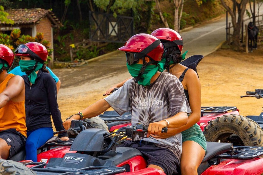 A group of people on a red ATV, wearing helmets and protective gear, on a dirt road with vegetation and a house in the background. 2
