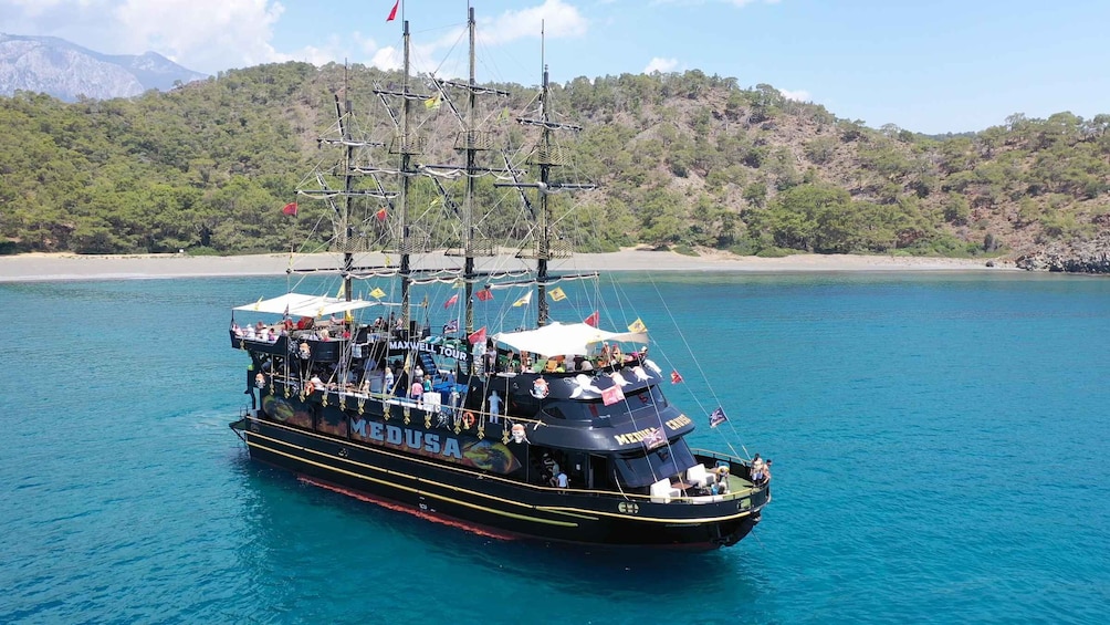 A large, black ship named 'MEDUSA' with multiple masts and passengers, sailing on clear blue waters with a forested coastline in the background. 1