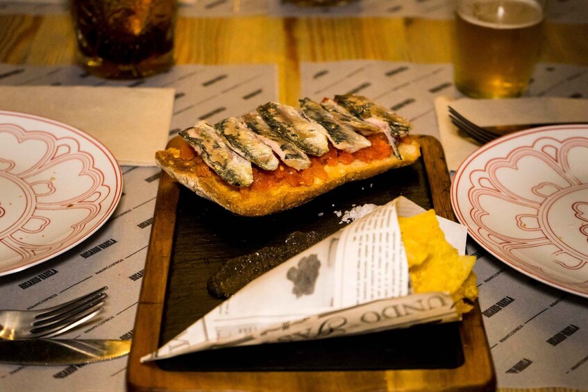 A wooden serving board with a dish of bread topped with anchovies and tomato sauce, a folded napkin, and a fork on a table with patterned placemats. 1