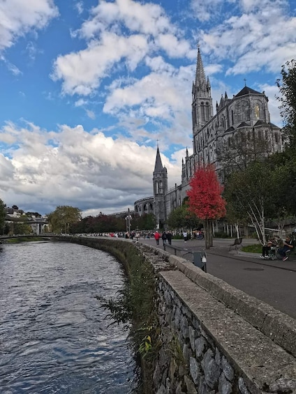 A river with a stone wall, a red tree, and a large cathedral with a tall spire. 1