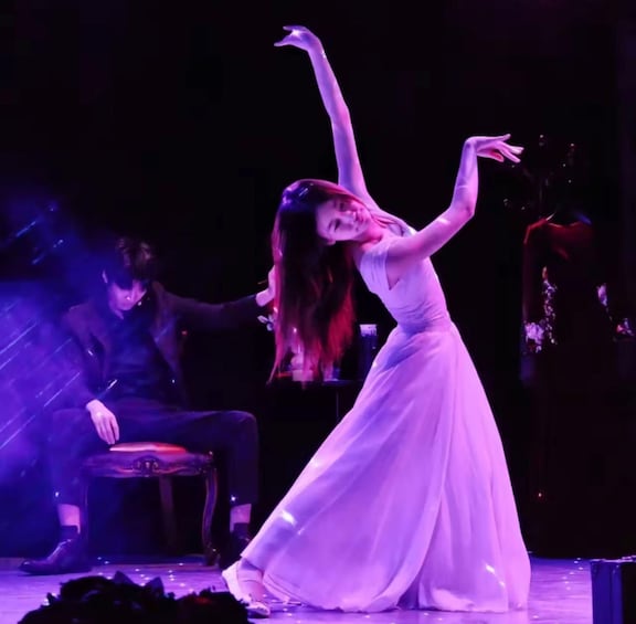 A person in a white dress performing on stage with a piano and another person in the background. 1