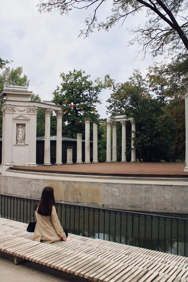 A woman sitting on a wooden bench by a canal with classical columns and statues in the background. 4