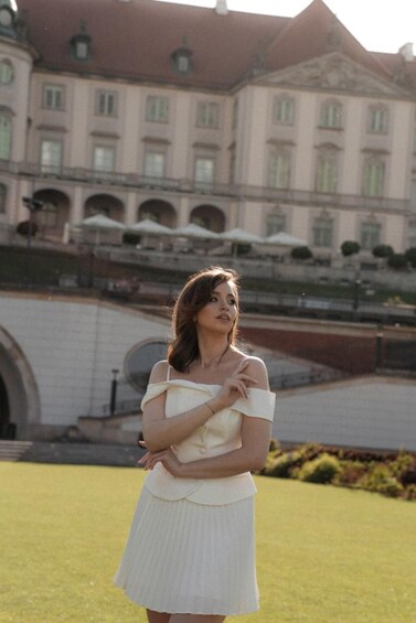 A woman in a white dress standing in a grassy area with a large building in the background. 1