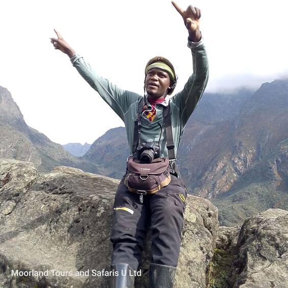 A person standing on a rocky outcrop with arms raised, wearing a jacket, pants, and boots, with a camera strapped to their chest. 2