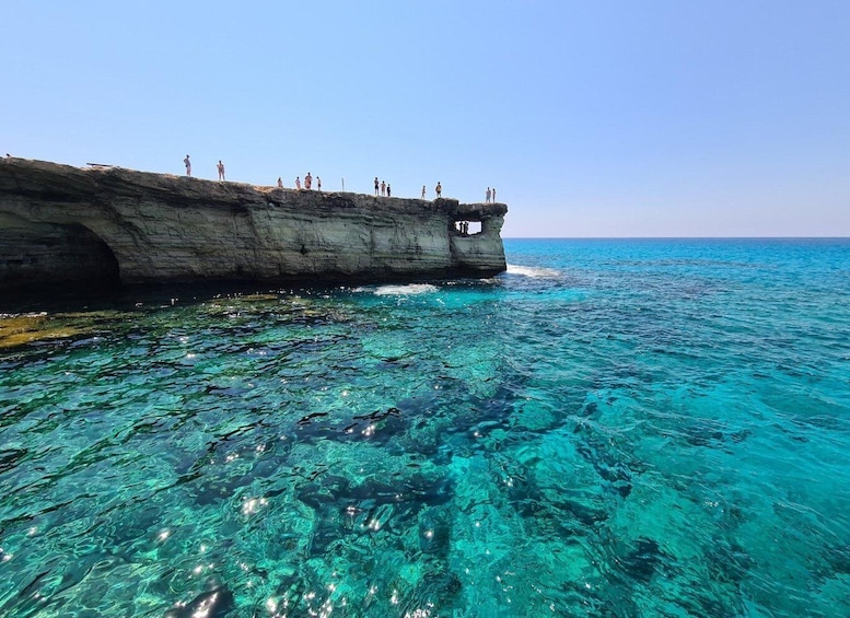A rocky coastline with clear turquoise water and people standing on the rock formation. 3