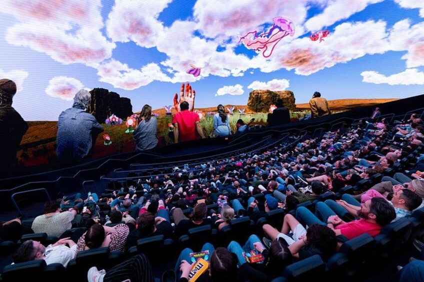 Un cinéma en plein air où un grand nombre de spectateurs sont assis en rangées, face à un écran qui diffuse des dessins animés et représente un paysage. 5