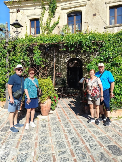 A group of people standing in front of a building with a brick pathway and a large potted plant. 1
