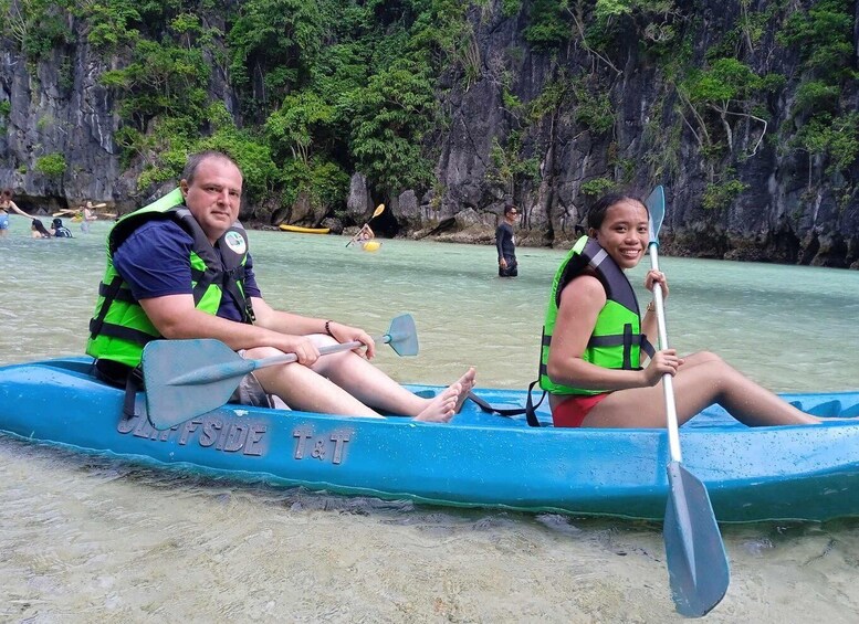 Two people in a blue canoe with 'SOUTHERN TRADE T&T' written on it, paddling on a calm river with a rocky cliff in the background. 3