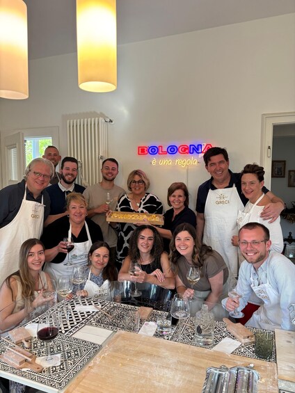 A group of people at a table with wine glasses, aprons, and a tray of food, with a neon sign reading 'BOLOGNA è una regola.' 3