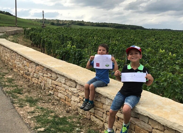Two children sitting on a stone wall in a vineyard, holding certificates. 2