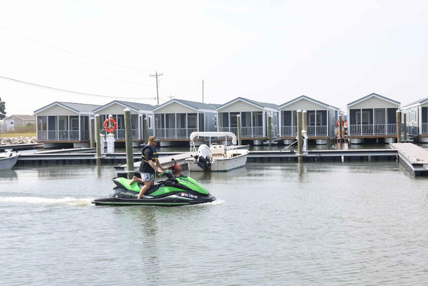 A person riding a green jet ski on calm water, with a row of houses in the background. 3