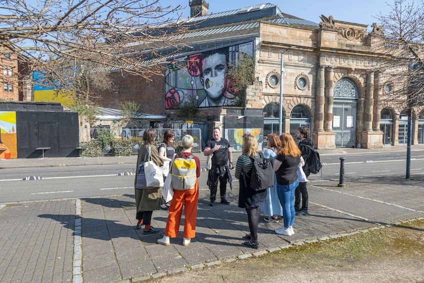 A group of people standing on a sidewalk, facing a large mural on a building. 5