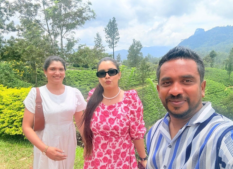 Three people standing in a lush green landscape with tea plantations and mountains in the background. 3